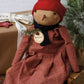 A rustic handmade Christmas doll wearing a red knit hat and a red-and-white gingham dress, sitting on a small wooden box beside a wrapped gift and a frosted green tree.
