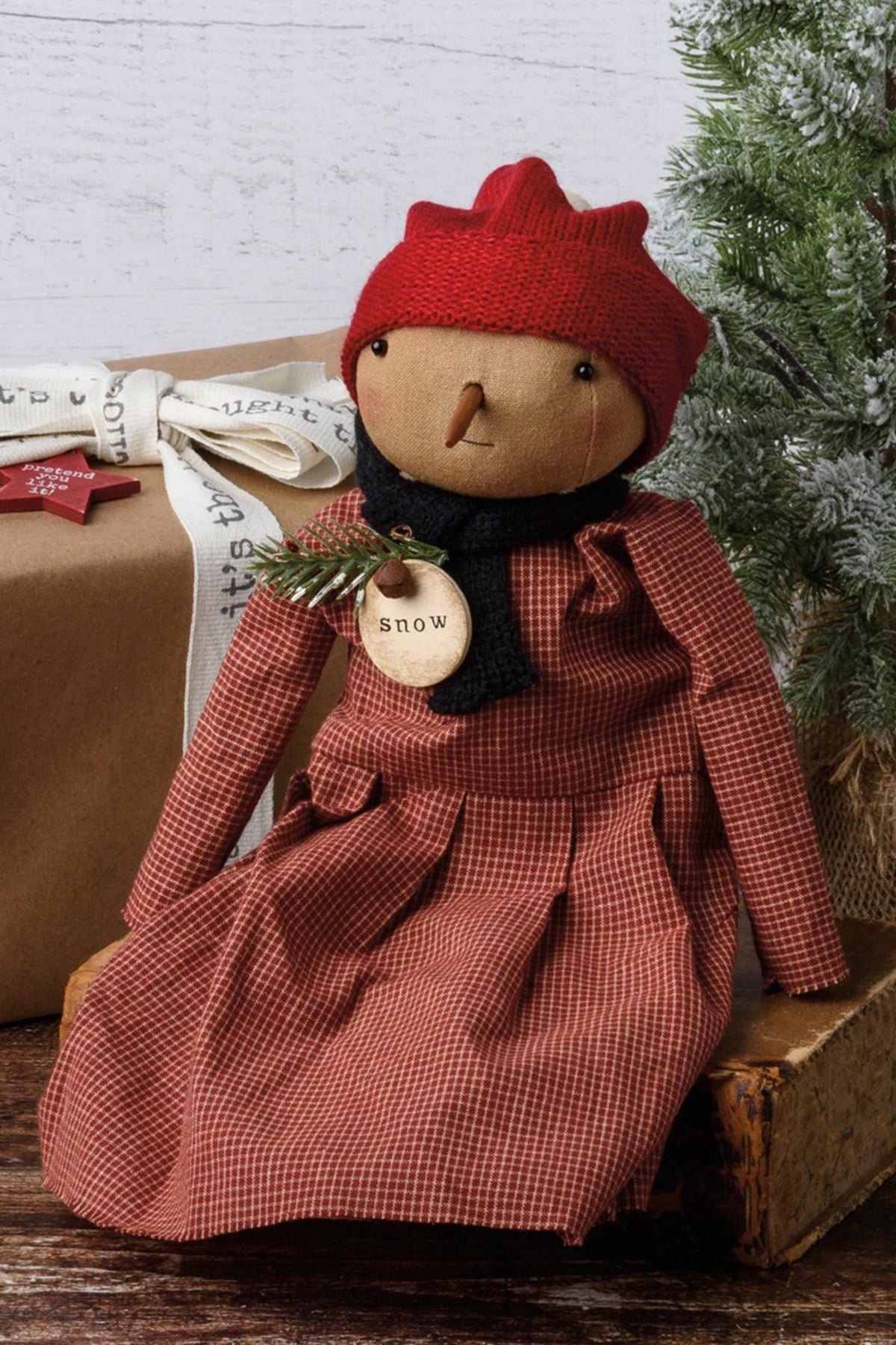 A rustic handmade Christmas doll wearing a red knit hat and a red-and-white gingham dress, sitting on a small wooden box beside a wrapped gift and a frosted green tree.