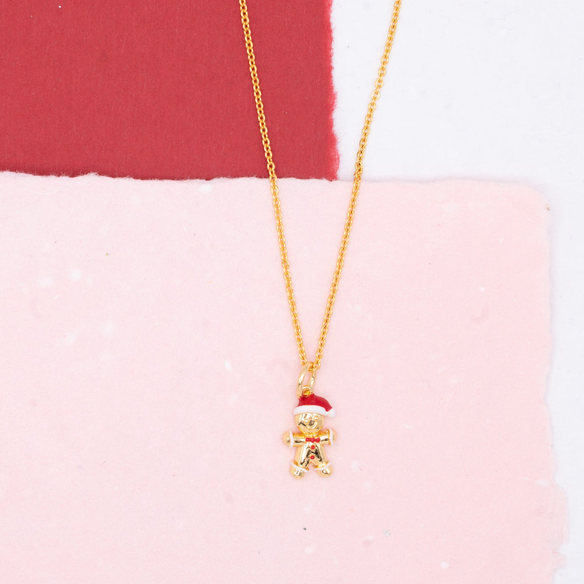 A gold chain necklace with a small gingerbread man pendant wearing a red Santa hat, displayed on a split pink and red background.