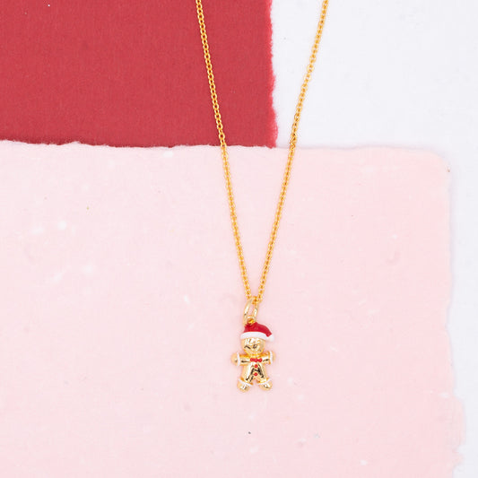 A gold chain necklace with a small gingerbread man pendant wearing a red Santa hat, displayed on a split pink and red background.