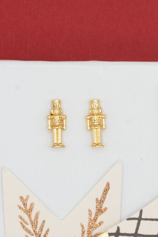 Gold-toned nutcracker stud earrings displayed on a white card alongside decorative star-shaped ornaments, with a red and white background.
