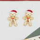 A pair of small gold-toned gingerbread person stud earrings with red Santa hats, displayed on a white gift box next to decorative star-shaped paper cutouts against a red background.