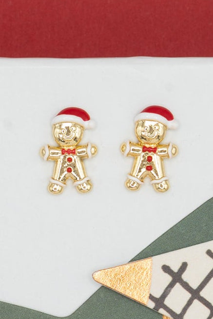 A pair of small gold-toned gingerbread person stud earrings with red Santa hats, displayed on a white gift box next to decorative star-shaped paper cutouts against a red background.
