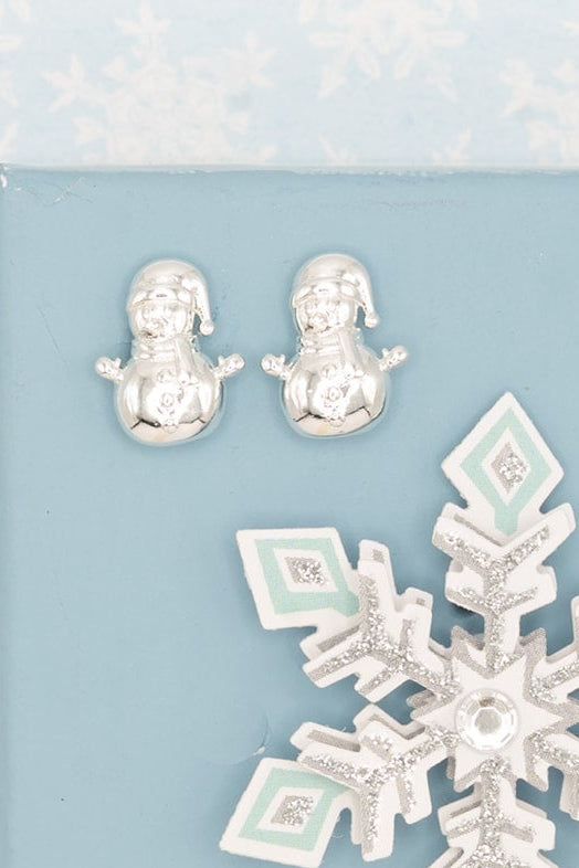A light blue rectangular jewelry display card or tray on a pale blue snowflake-patterned background, holding three silver-toned jewelry pieces: a large snowflake charm or brooch in the center and two small snowman-shaped studs or charms near the top.