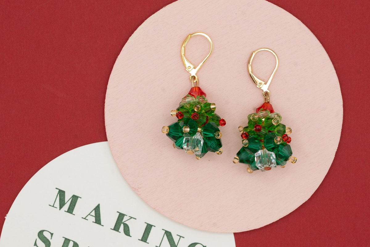 A pair of gold leverback earrings shaped like Christmas trees, featuring green faceted beads, red accents, and a clear crystal, displayed on a pink and white circular card over a dark red background.