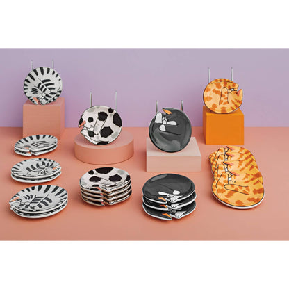 A styled product display of multiple ceramic plates with animal and abstract patterns, arranged on pastel-colored pedestals against a split lavender and peach background.