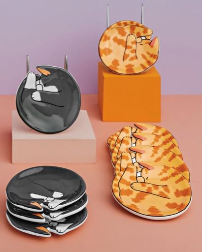 A set of ceramic plates featuring a whimsical cartoon cat design, displayed on minimalist pedestals against a pastel purple and peach background. The plates include a black cat curled up and a yellow-orange tabby cat with an open mouth.